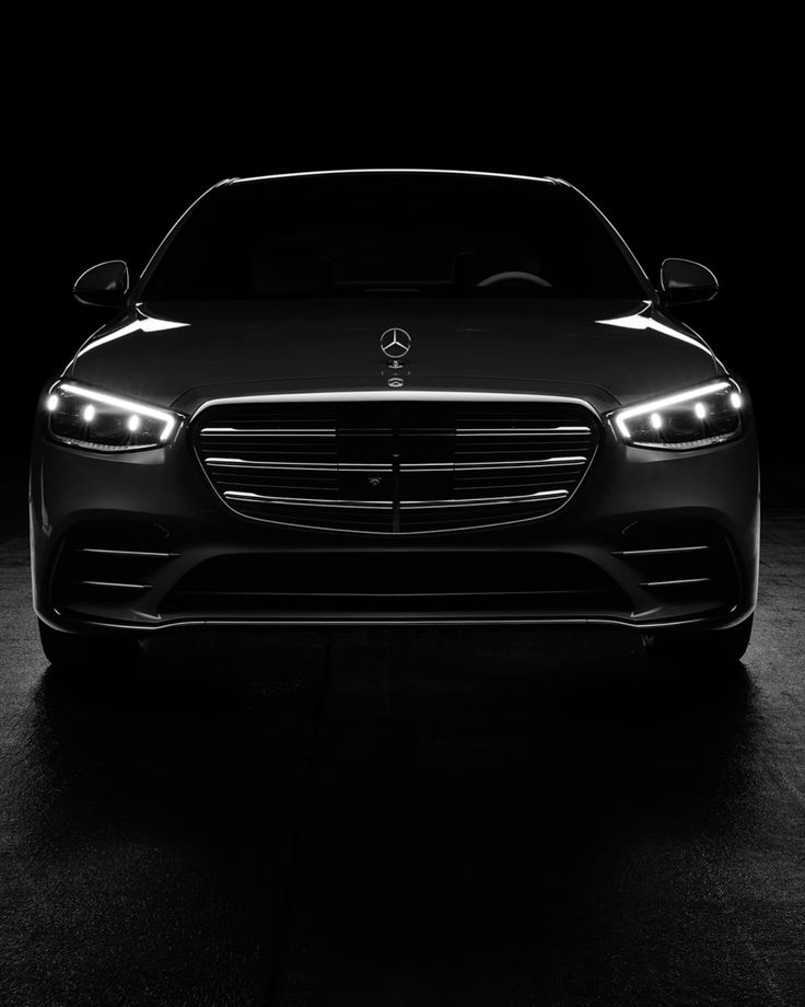 Frontansicht eines dunklen Mercedes-Benz mit eingeschalteten Scheinwerfern vor schwarzem Hintergrund.