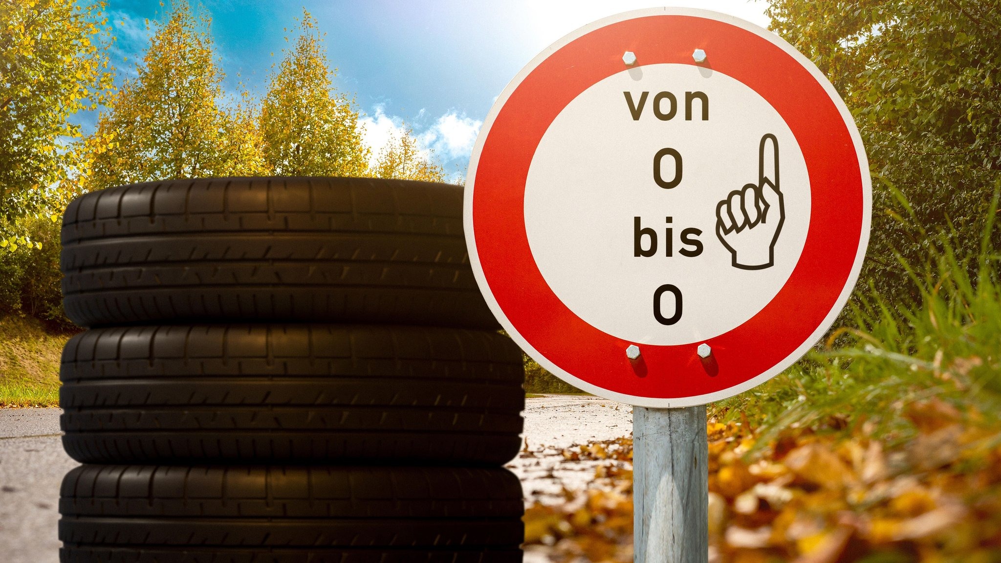 Gestapelte Autoreifen neben einem Schild mit der Aufschrift „von O bis O“ in einer Herbstlandschaft.