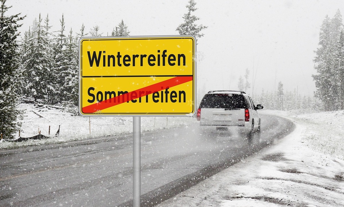 Verschneite Straße mit einem Auto und einem gelben Schild „Winterreifen“ mit durchgestrichenem „Sommerreifen“.